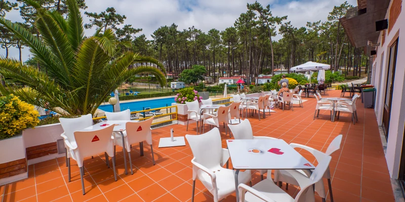 White chairs and tables arranged on a tiled terrace overlooking a pool and pine forest, at Valado