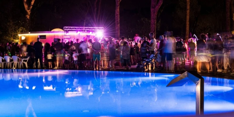 A crowd gathers around a brightly-lit pool at night during a lively event, surrounded by tall trees at Cieloverde