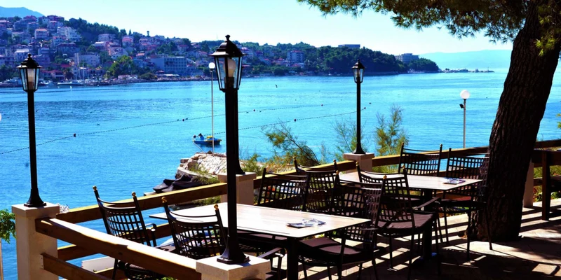 Tables and chairs sit empty on a terrace overlooking a serene, sunlit seaside town and calm blue water at Stobreč Split