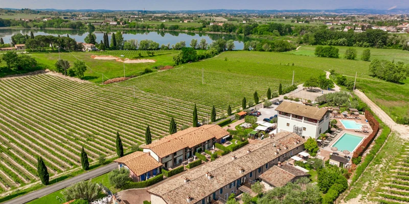 Vineyard rows stretch beside farmhouse buildings and two swimming pools, overlooking a tranquil lake surrounded by lush greenery at La Bertoletta Village apartments
