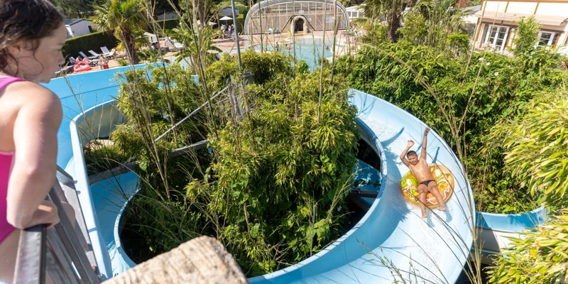 Child sliding down blue water slide, surrounded by lush greenery and pool area with sunbathers in the background at La Côte de Nacre
