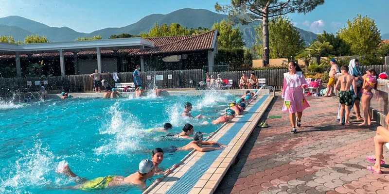People swimming and splashing in an outdoor pool surrounded by mountains and trees, with others watching or standing nearby at Del Sole