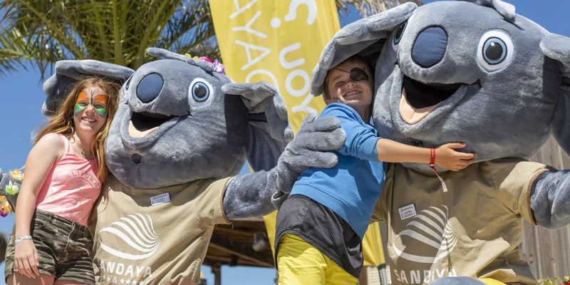 Two children posing with giant koala mascots, smiling under a palm tree and a yellow banner at Cap Sud