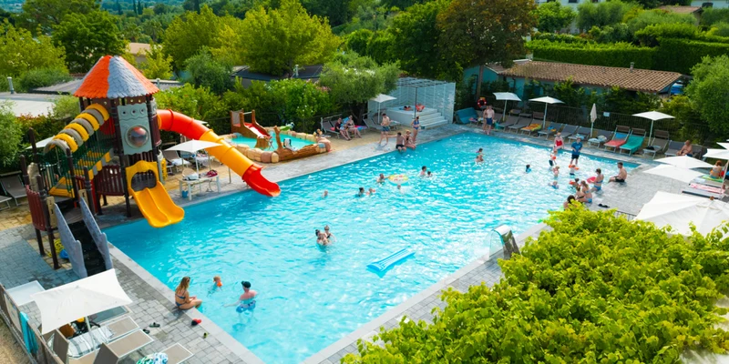 People swimming in a pool with slides and play area, surrounded by trees and sun loungers at Vacanze Glamping Boutique