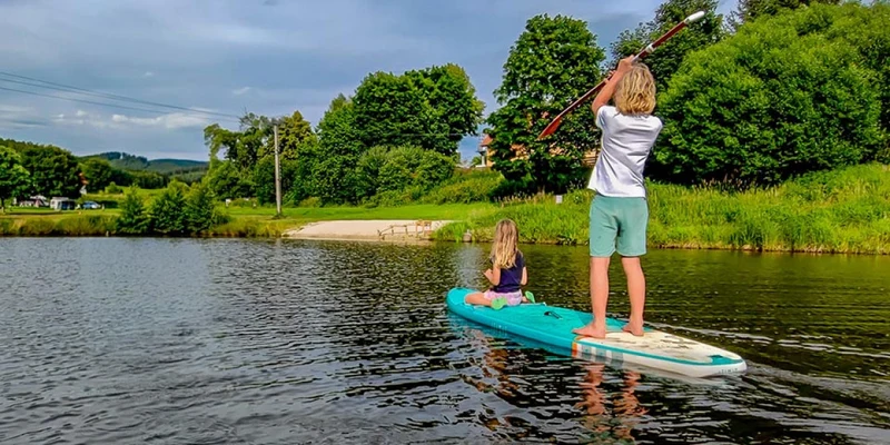Two children are paddleboarding on a calm lake, surrounded by lush green trees and a sandy shore at Chvalsiny