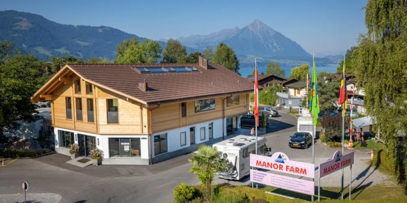 A wooden building with skylights sits beside parked vehicles, surrounded by colorful flags and mountains at Manor Farm