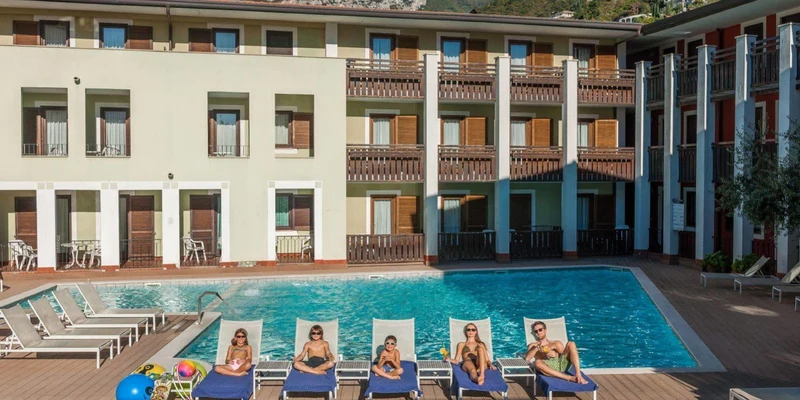 Six people sunbathe on lounge chairs beside a swimming pool, next to a three-story hotel with mountain backdrop at Holiday Park La Vela