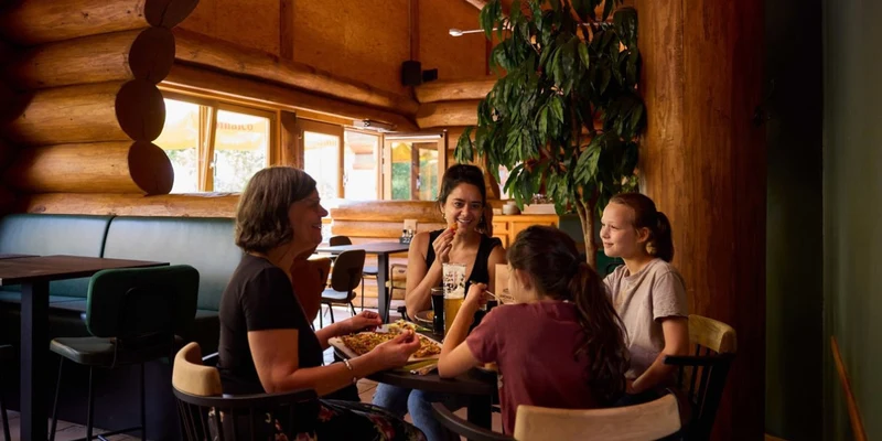People eating and chatting inside a wooden log cabin restaurant with warm lighting and green plants at Laacher See