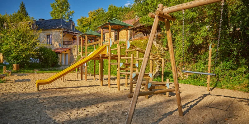 Playground with swings and a yellow slide, surrounded by trees and rustic buildings at Aqua Viva