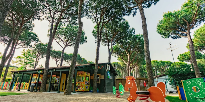 Toy horse on a spring stands in a sandy playground surrounded by tall trees and a building at Camping Village Florenz