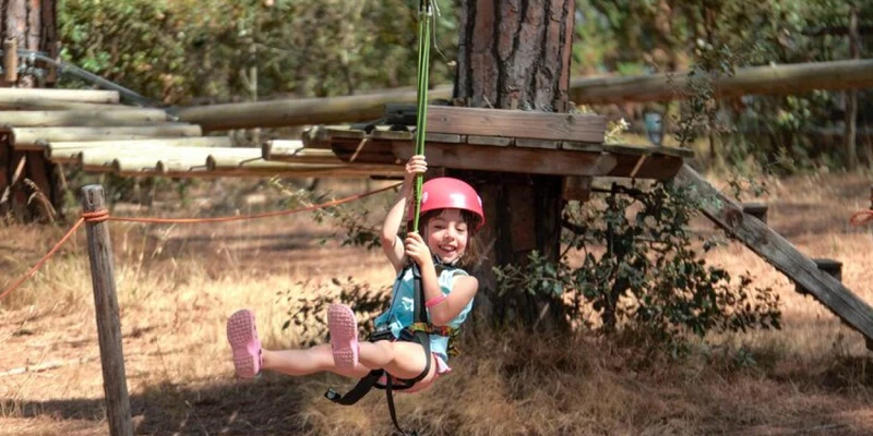 Child hanging on zip line wearing safety gear in wooded area with trees and wooden platforms at Le Marze