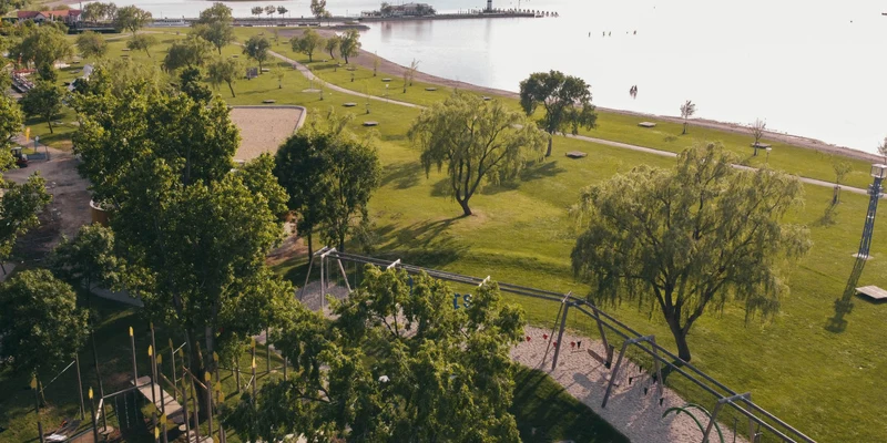 Playground with swings situated on sandy area, surrounded by lush green parkland near a scenic waterfront at Podersdorf