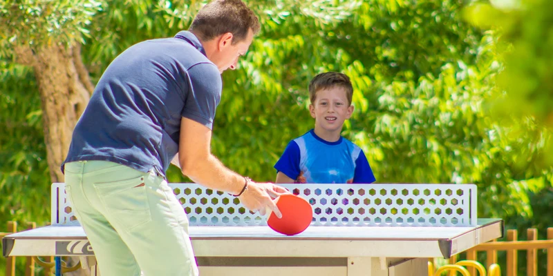 Two individuals playing table tennis amidst lush green trees at Ametlla