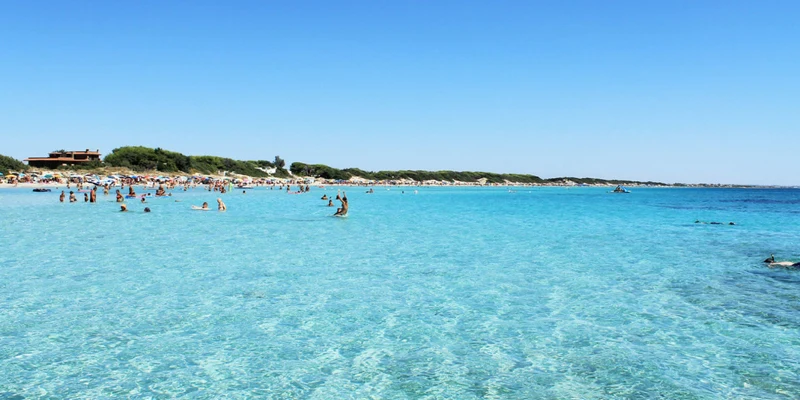 Swimmers enjoying clear, blue water on a sunny day; crowded beach with sunbathers in the background at Porto Cesareo Camping