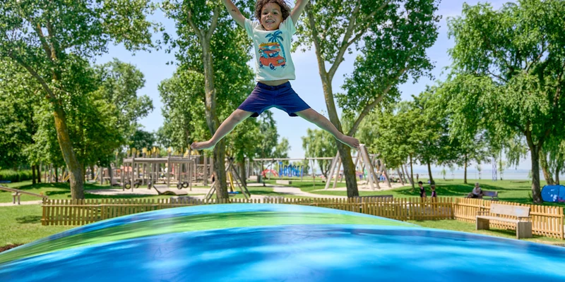 Child joyfully jumps on a colorful inflatable pad, surrounded by trees and playground equipment at Podersdorf