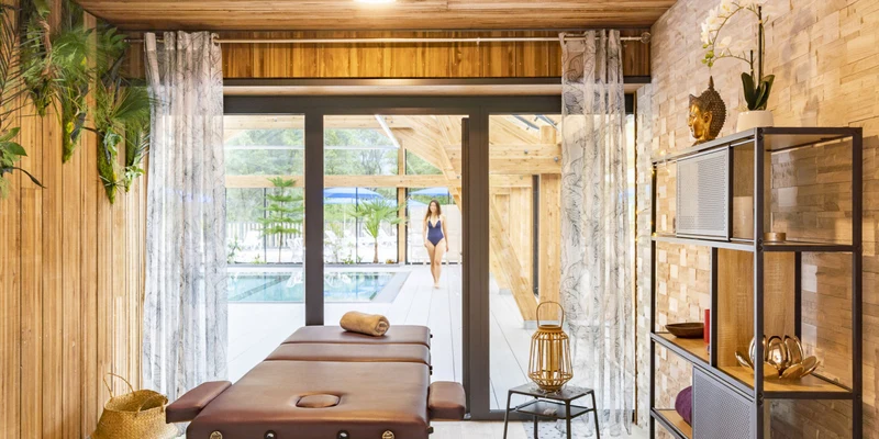 Massage table stands in a cozy, wooden treatment room overlooking a pool area with a person walking at La Nublière