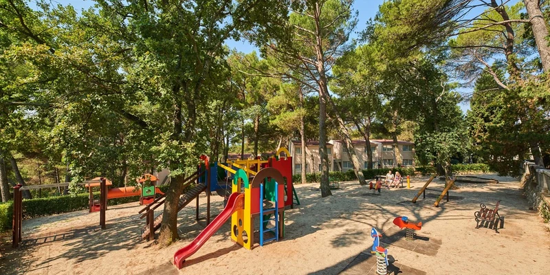 Colorful playground equipment stands idle amidst tall, green trees in an outdoor recreational area at Bellevue Plava Laguna