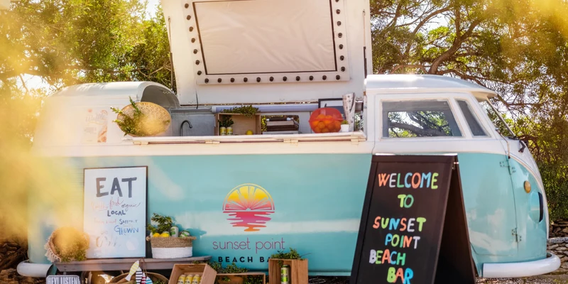 Blue retro van converted into a beach bar, surrounded by trees, displays fresh produce and welcoming signs at Waterman Beach Village