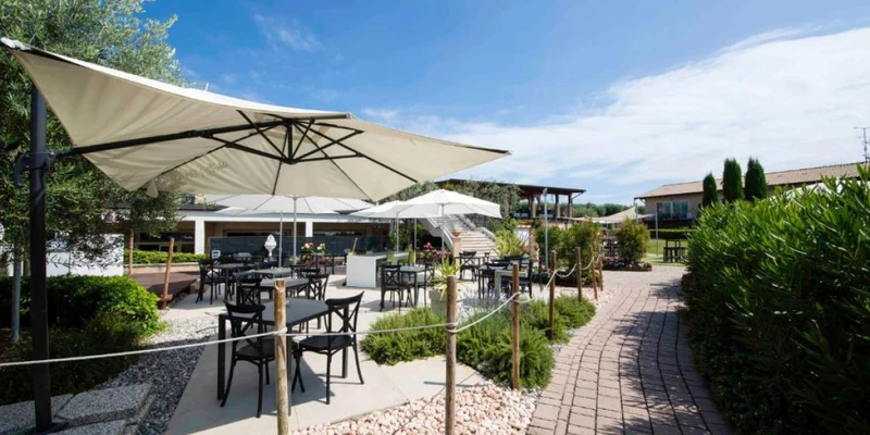 Outdoor patio with tables and chairs under large umbrellas, along a stone pathway, surrounded by greenery at Lake Garda Resort
