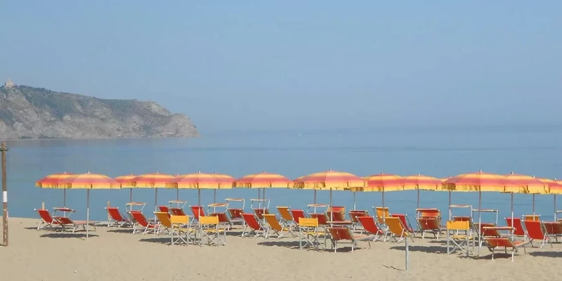 Yellow and orange beach loungers and umbrellas are lined up on a sandy beach with calm sea and rocky cliffs in the background at Tindari Village