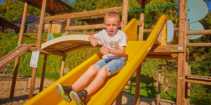 Child sliding down a yellow slide on a wooden play structure, surrounded by greenery and sand at Aqua Viva