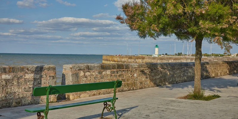 Green bench sits by a stone seawall, overlooking the ocean with a lighthouse in the distance, under a tree at Marvilla Parks La Côte Sauvage