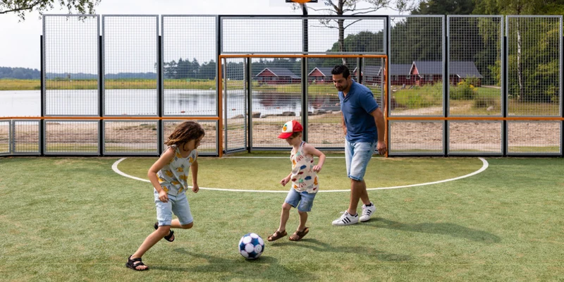 Three people play soccer on an outdoor field near a lake, with tall netted fences surrounding the area at de Flaasbloem