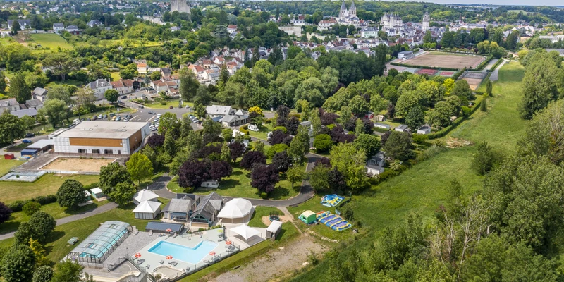 A swimming pool with adjacent tents in a tree-filled campground near buildings and a city with a church tower; aerial view at La Citadelle
