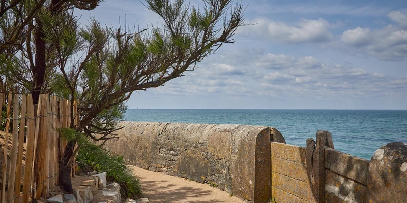 Stone pathway curves beside a wooden fence and tree, leading to a tranquil sea view at Marvilla Parks La Côte Sauvage