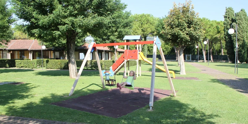 Child swinging on playground swing set; slide and climbing structure surrounded by green grass and trees at Front Lake Resort Le Corti del Lago