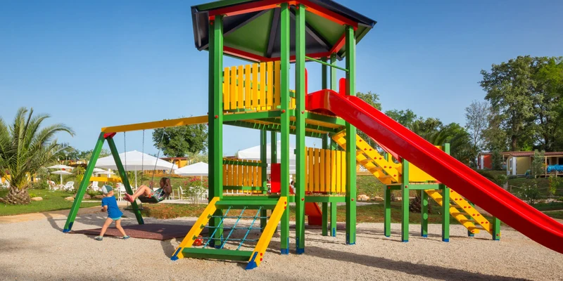 Colorful playground with slide and swings, children playing joyfully, surrounded by trees and picnic tables at Aminess Maravea Camping Resort