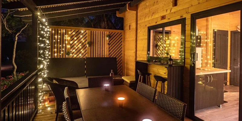 Wooden patio illuminated with string lights, placed next to a cozy seating area and dining table with candles at Orangeraie