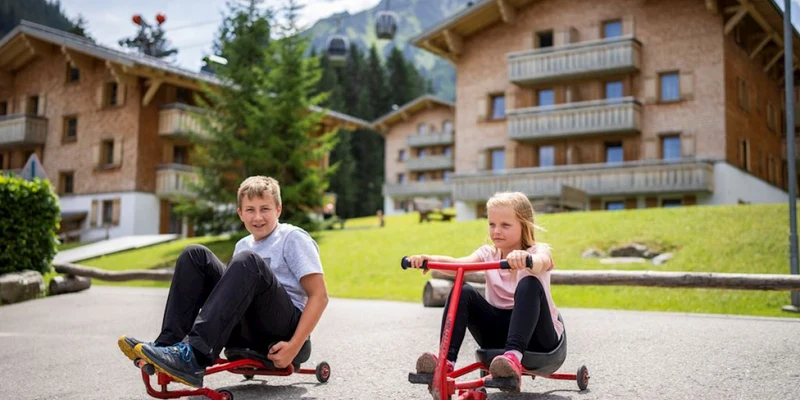 Two children ride small red trikes on a paved path near wooden chalets and mountains at Landal Hochmontafon