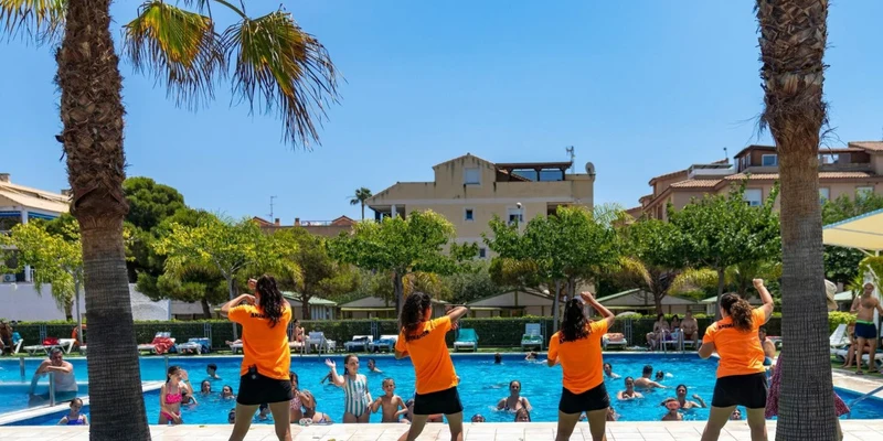 Instructors in orange shirts lead an outdoor exercise session by a pool, surrounded by people and palm trees at Valencia