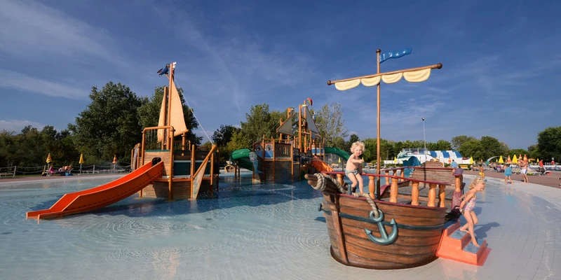 Wooden pirate ship play structure with slides and children playing in a shallow pool, surrounded by trees and lounge chairs at Pra delle Torri