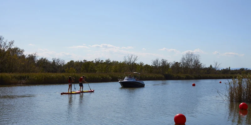 People paddleboarding and boating on a calm river surrounded by trees and red buoys at Nautic Almata