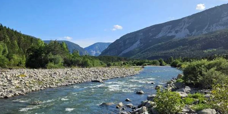 River flowing past rocky banks with bushes and a forest against a backdrop of mountains under a clear blue sky at Paradis Les Relarguiers