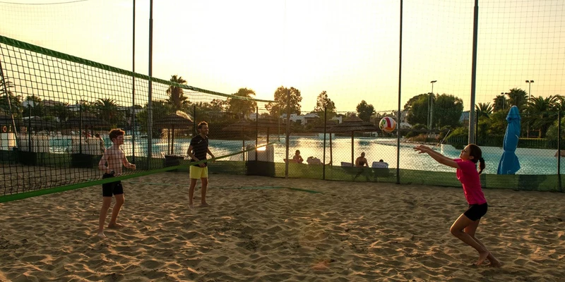 Three people playing beach volleyball at sunset on a sand court near a swimming pool, surrounded by trees at I Turchesi Club Village