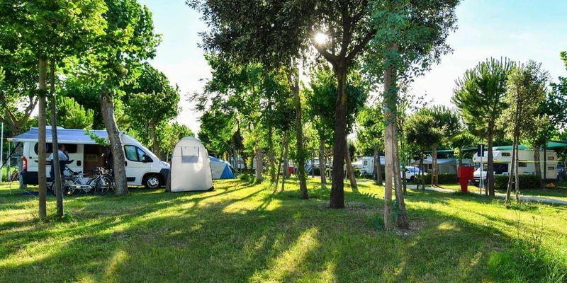 Campervan with canopy and bikes parked amid trees and tents in a sunny grassy camping area at Riva Nuova