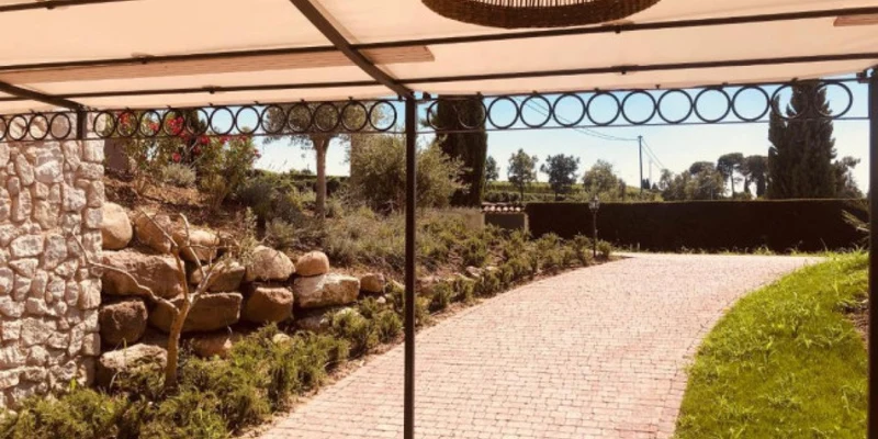 Covered patio overlooking a sunlit, winding brick path with stone wall and green shrubs at La Bertoletta Village apartments