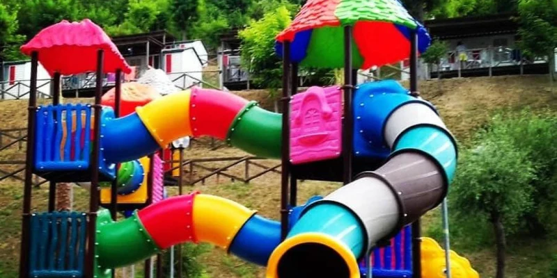 Colorful playground slides intertwine around a tower, set in a green, hillside park with cabins in the background at Camping Village Mirage