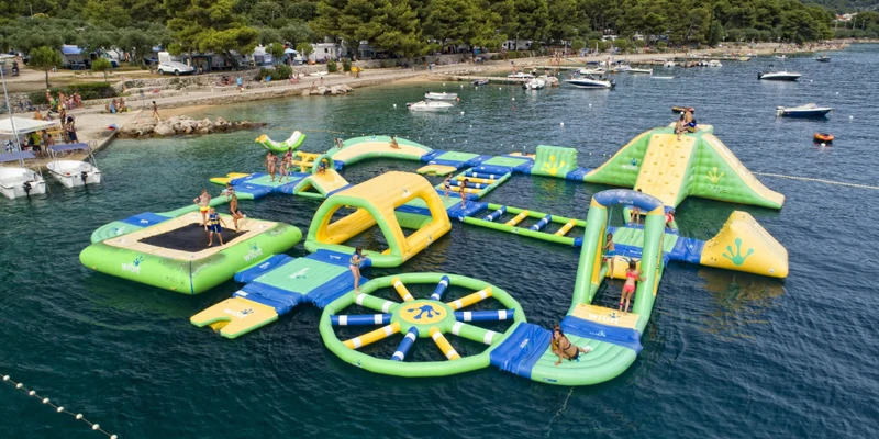 Inflatable water park with people playing on various structures, floating in a picturesque lake surrounded by trees at Kovacine