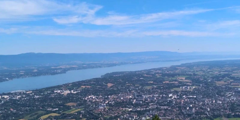A sprawling cityscape borders a large, serene lake, surrounded by distant mountains under a clear blue sky at Relais du Léman