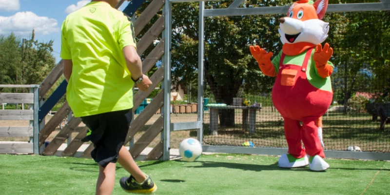 Child in neon shirt kicks ball towards large fox mascot on green field with wooden structure at Le Coin Tranquille