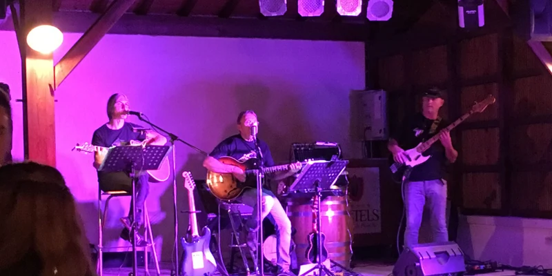 Three musicians perform on a small indoor stage with instruments and microphones under purple lighting at Le Camp de Florence