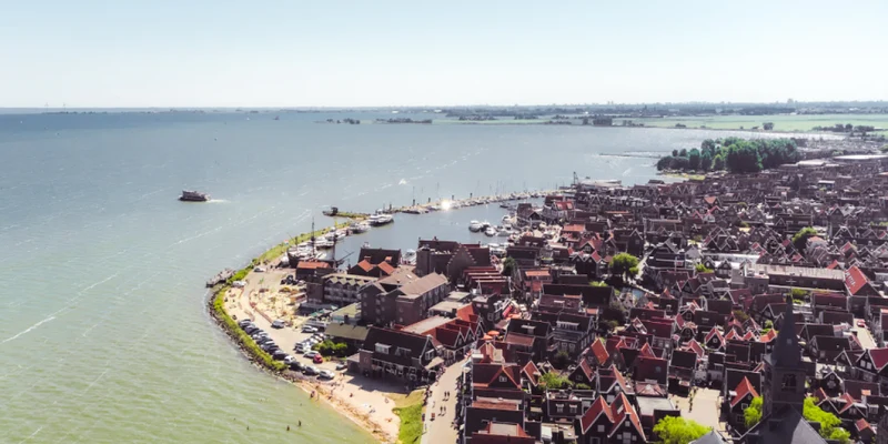 A coastal town with red-roofed buildings and a harbor filled with boats, surrounded by a shimmering sea and green land at Park Wijdenes
