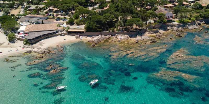 Boats float on clear turquoise water near rocky shoreline and sandy beach, surrounded by trees and buildings at U Livanti