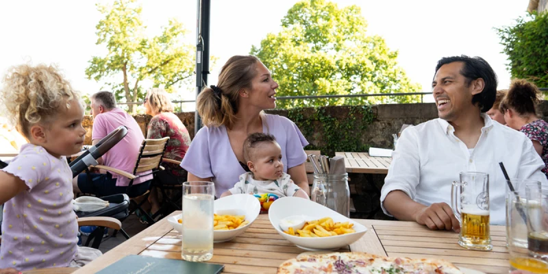 Family dining outdoors, talking and enjoying drinks and pizza, under an umbrella with trees in the background at Ferienresort Bad Bentheim