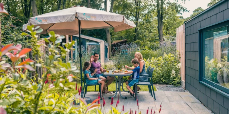 Family eating at an outdoor table under an umbrella, surrounded by greenery and cabins at Europarcs De Utrechtse Heuvelrug