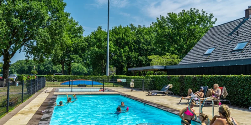 People swim and relax around a swimming pool, surrounded by trees and a house with a steep roof at Europarcs Reestervallei